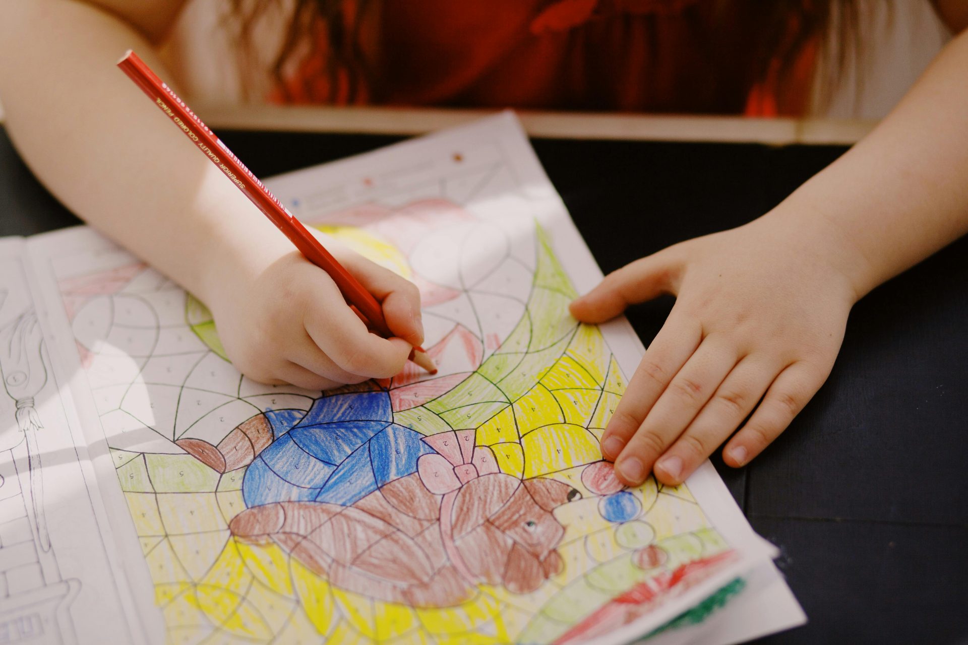 Primo piano delle mani di un bambino che usa una matita colorata rossa per riempire una pagina di un libro da colorare numerato con un cane cartone